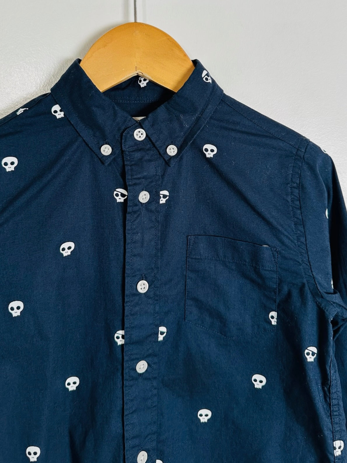 A dark navy blue button-down shirt with a white skull pattern, hanging on a wooden hanger against a plain light gray wall.