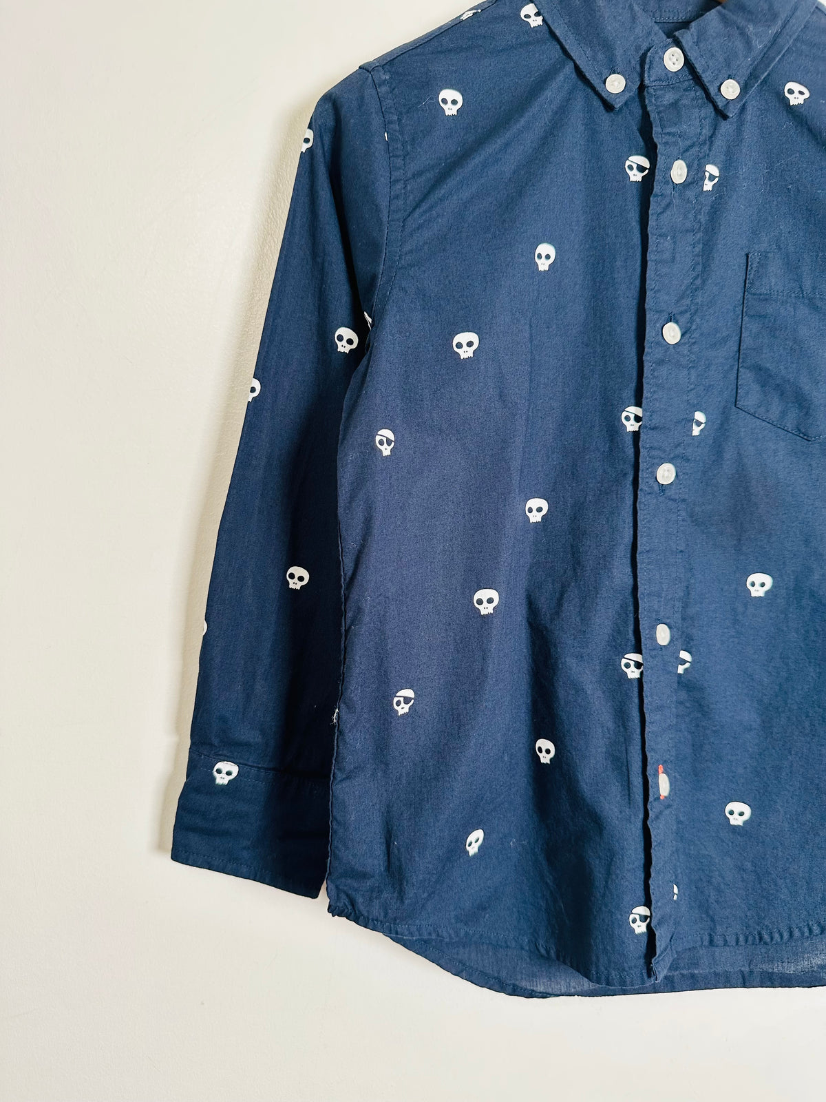 A dark blue long-sleeve button-up shirt with a repeating white skull pattern, shown hanging against a plain off-white wall.
