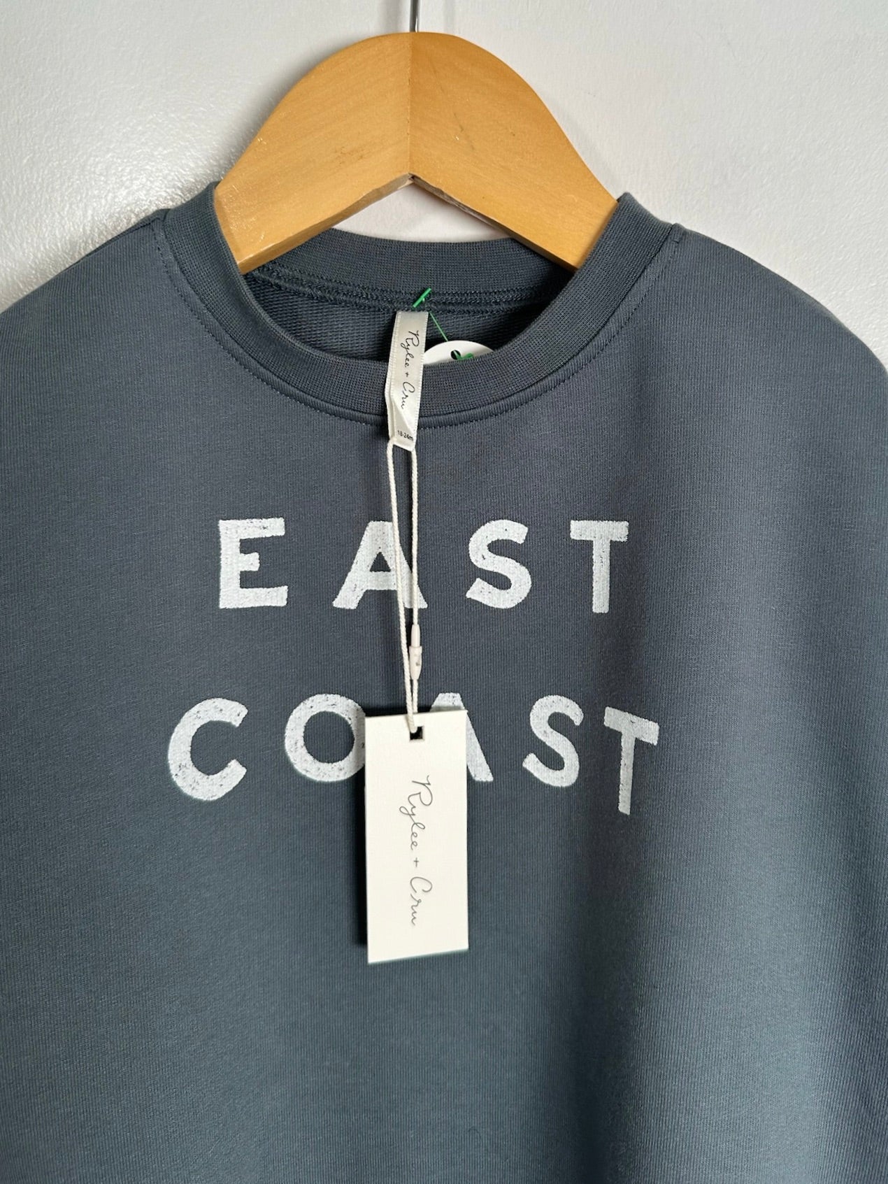 A dark slate gray crewneck sweatshirt hanging on a wooden hanger against a light gray wall. The sweatshirt features the words 'EAST COAST' printed in a distressed white font across the chest, with a white hangtag attached to the collar.