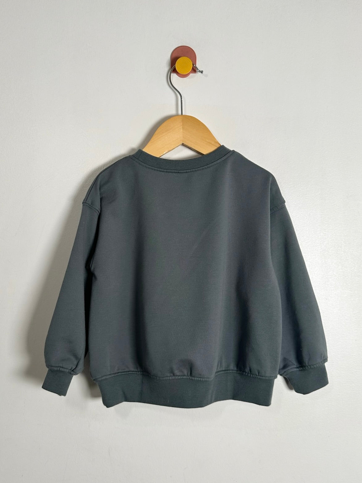A dark gray crewneck sweatshirt hanging on a wooden hanger against a plain off-white wall.