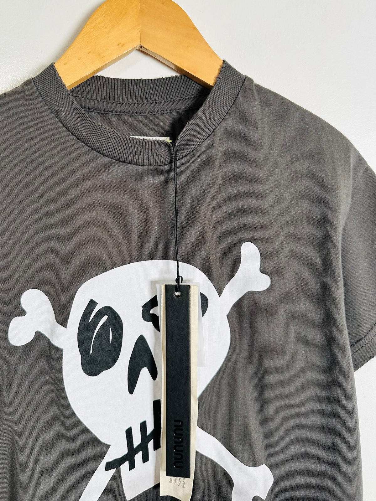 A dark gray crewneck t-shirt on a wooden hanger, featuring a large white skull and crossbones graphic with black details on the front. A black hangtag with the brand name 'Nununu' is attached to the collar.