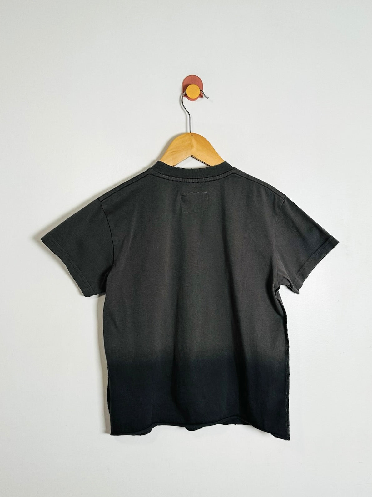 A black short-sleeve t-shirt with a dark-to-light gradient fade at the hem, hanging on a wooden hanger against a plain white wall.