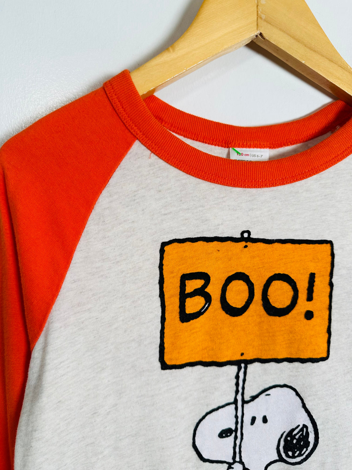 A close-up of a raglan-style long-sleeve shirt hanging on a wooden hanger against a white wall. The shirt has bright orange sleeves and collar, a light heather gray body, and a graphic of a cartoon sign reading 'BOO!' with a white dog character below.