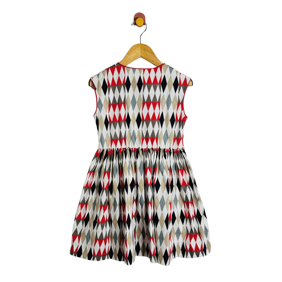 A sleeveless children's dress with a geometric diamond pattern in red, black, gray, and beige, hanging on a wooden hanger against a plain white background.