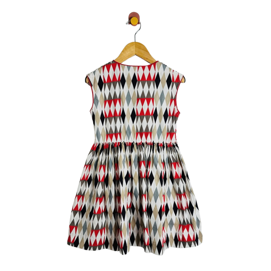 A sleeveless children's dress with a geometric diamond pattern in red, black, gray, and beige, hanging on a wooden hanger against a plain white background.