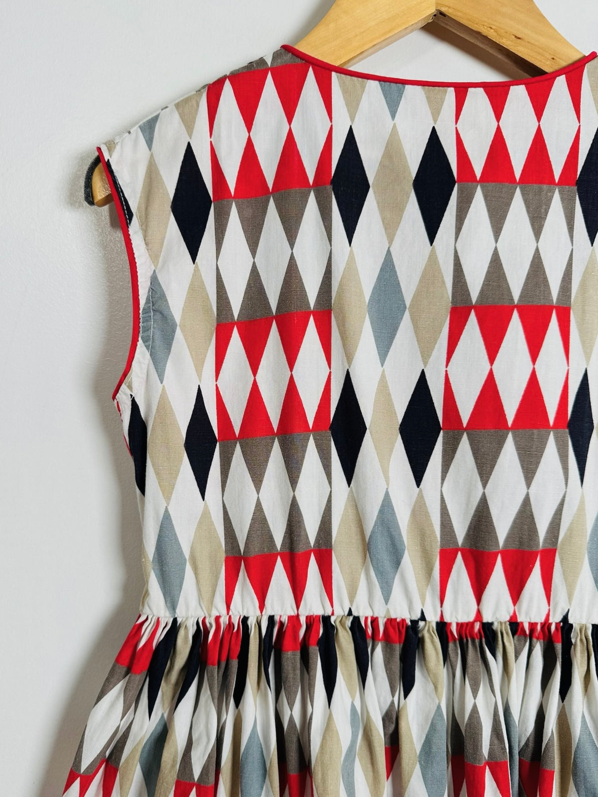 A sleeveless dress with a geometric harlequin diamond pattern in red, black, beige, gray, and white, hanging on a wooden hanger against a plain white wall.