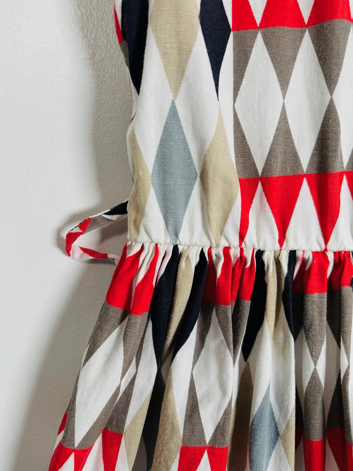 Close-up of a sleeveless dress with a geometric harlequin diamond pattern in red, beige, gray, black, and white, featuring a gathered waist seam and a side tie detail.