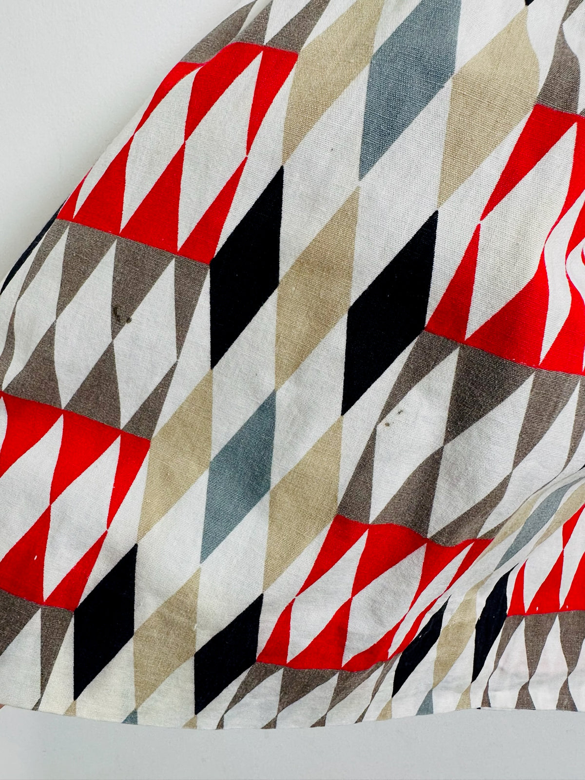 A close-up of a draped fabric with a geometric pattern of red, black, beige, gray, and light blue triangles on a white background.