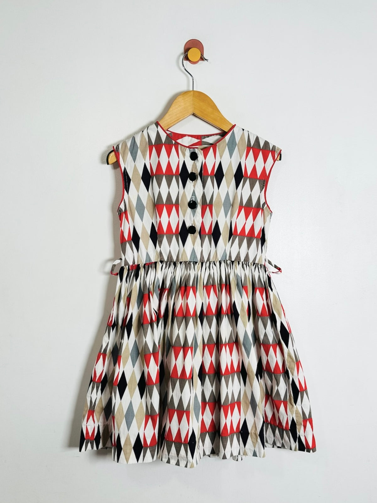 A sleeveless children's dress with a geometric harlequin diamond pattern in red, black, gray, and white, hanging on a wooden hanger against a plain light gray wall.