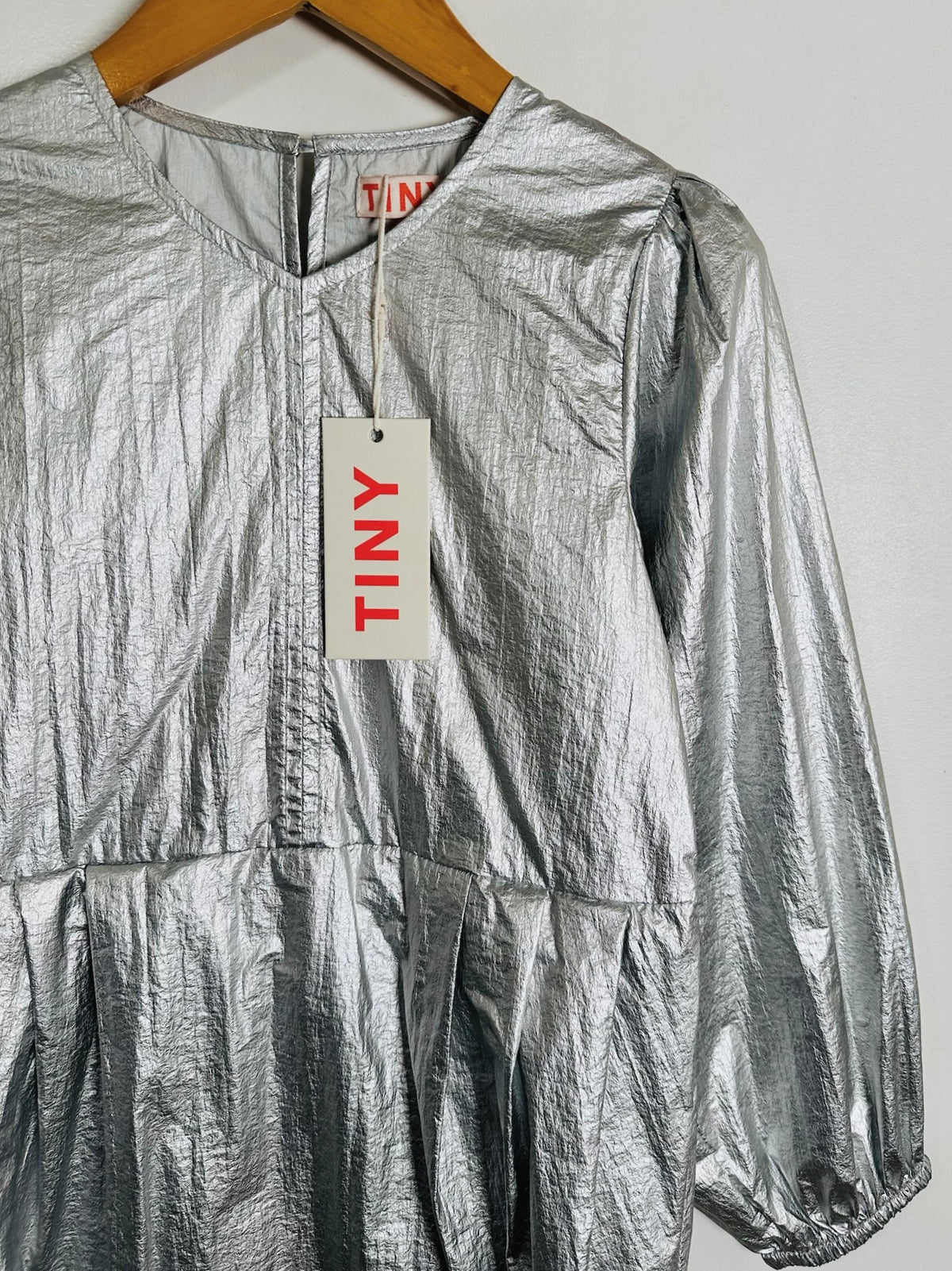 A metallic silver crinkled long-sleeve top hanging on a wooden hanger against a plain light gray wall, with a white paper tag reading TINY attached at the neckline.