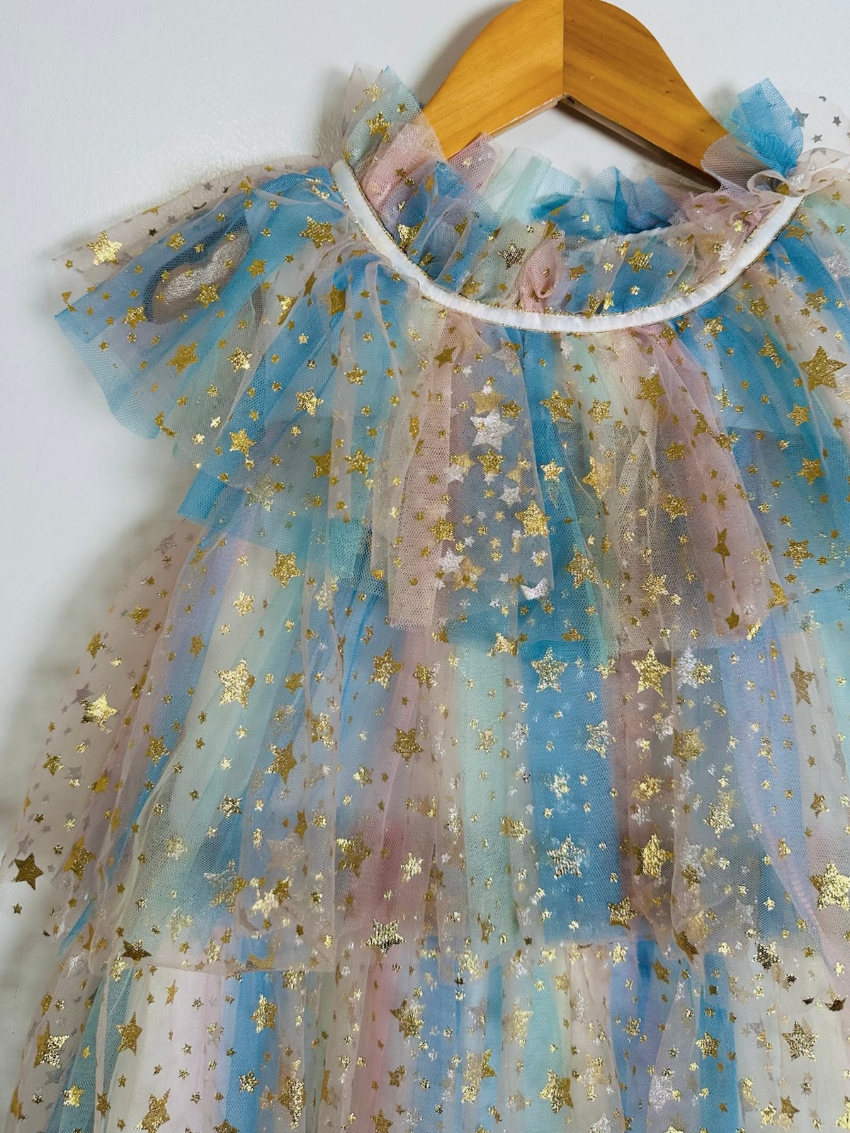 A tiered tulle dress for toddlers or girls, featuring a pastel blue, pink, and white gradient with scattered gold star prints, hanging on a wooden hanger against a plain white wall.