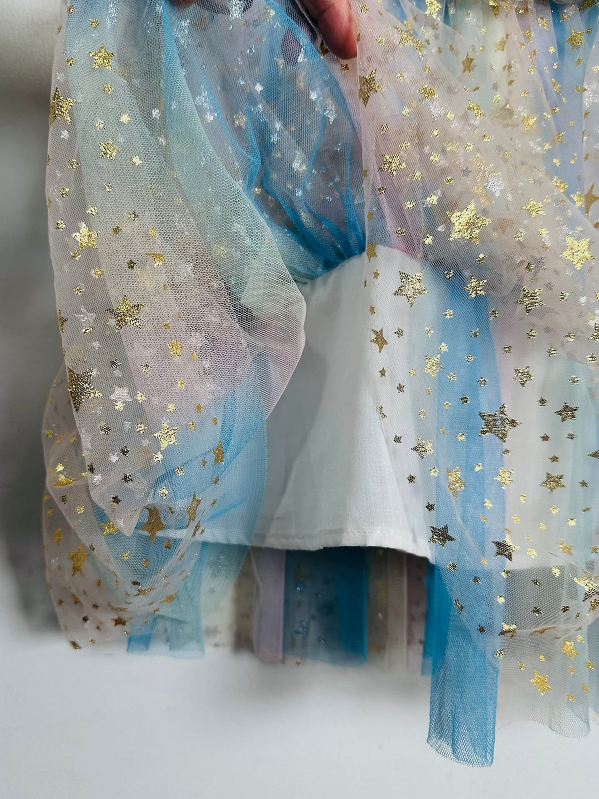 A close-up of a sheer tulle fabric with a pastel gradient of blue, purple, and white, decorated with scattered gold star confetti, held by a hand.