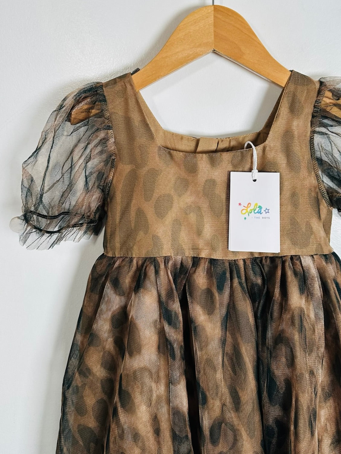 A girl's dress with a brown and black leopard print pattern, featuring sheer black tulle puff sleeves and a square neckline, hanging on a wooden hanger against a white wall. A white brand tag for 'Cute Little Star' is attached.
