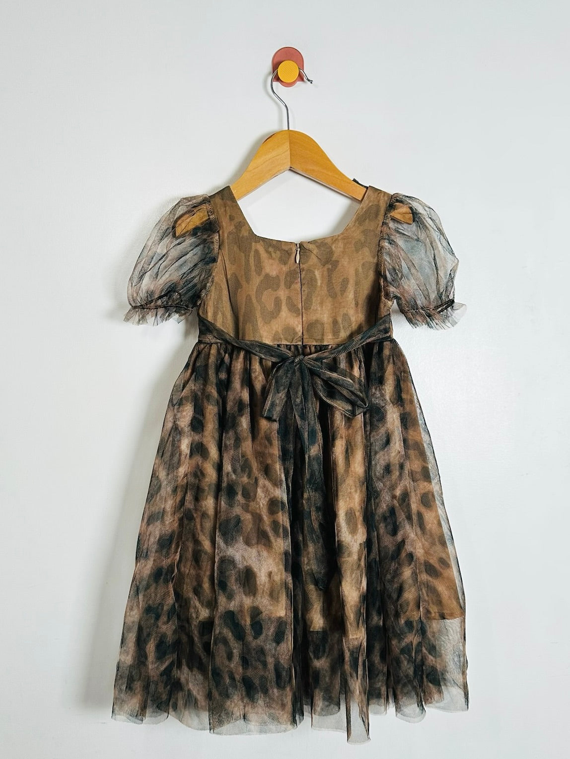 A children's dress with a brown and black leopard print pattern, featuring sheer puff sleeves, a square neckline, and a matching fabric tie belt at the waist, hanging on a wooden hanger against a plain white wall.