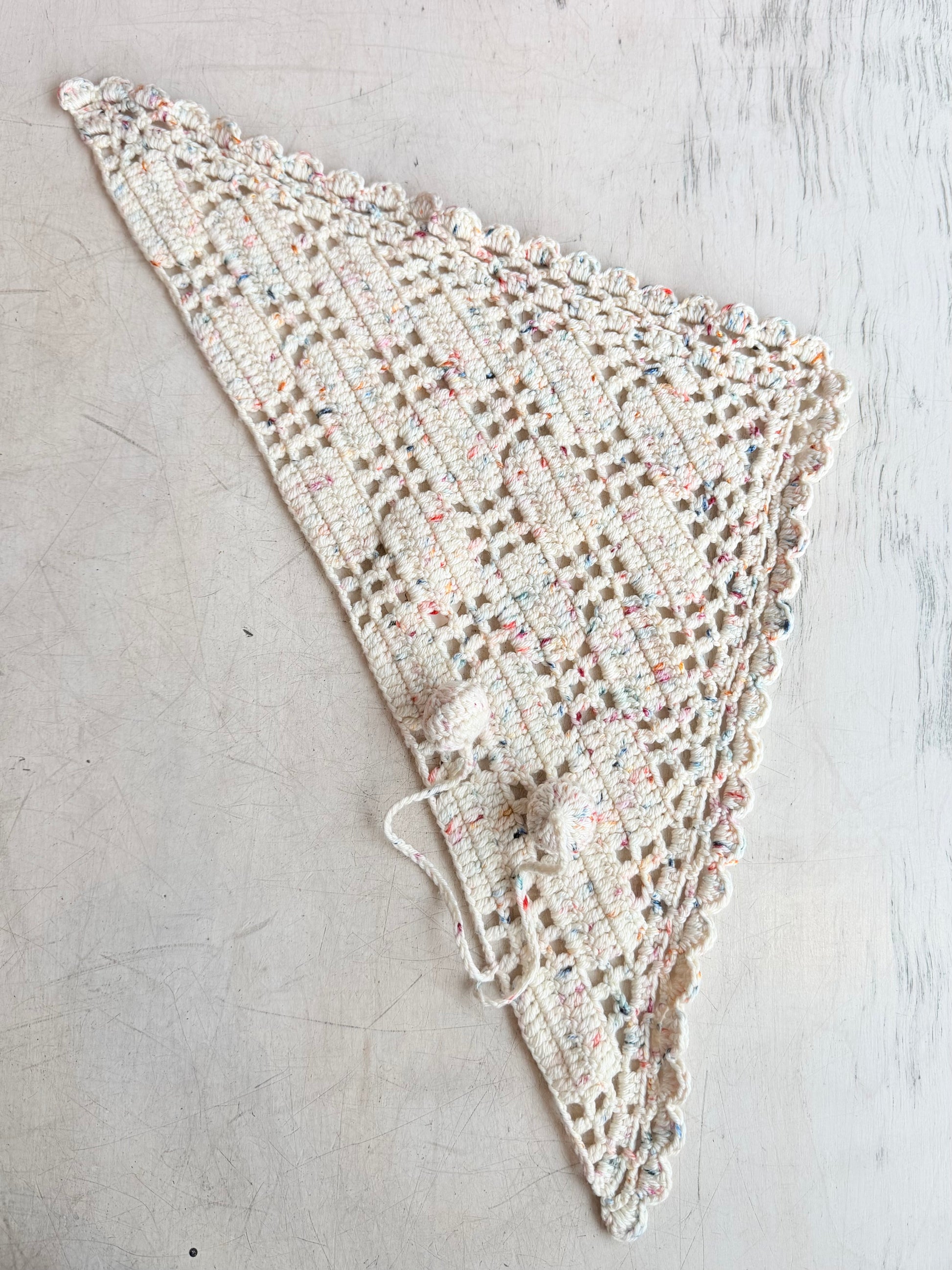A handmade triangular crochet shawl in cream and multicolor yarn, laid flat on a light gray textured surface.