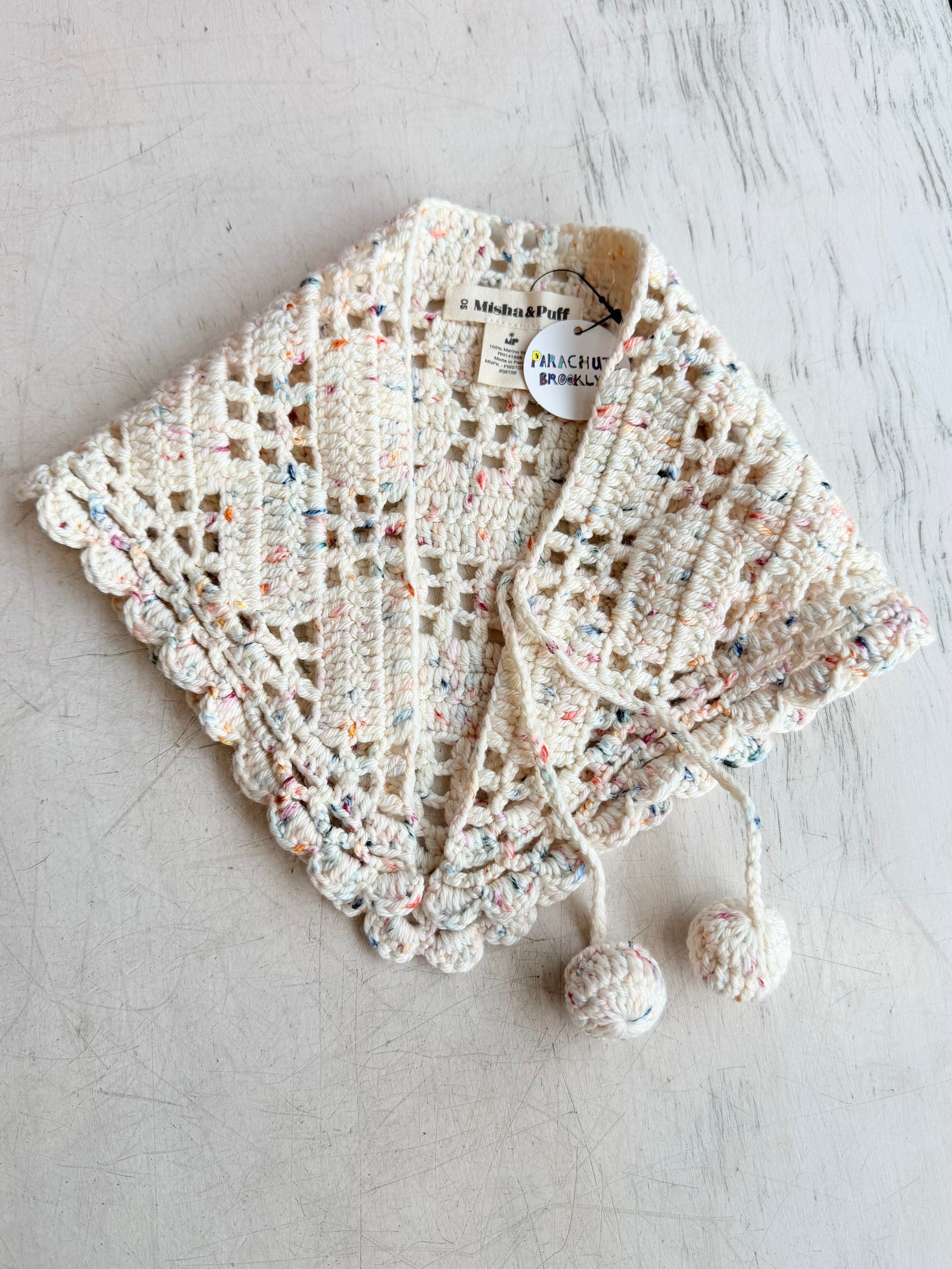 A handmade crochet shawl or wrap in cream with multicolored flecks, featuring a scalloped edge, drawstring closure, and two pom-poms, laid flat on a light textured surface. A brand label for 'A Minna & Nell' is visible inside the collar.