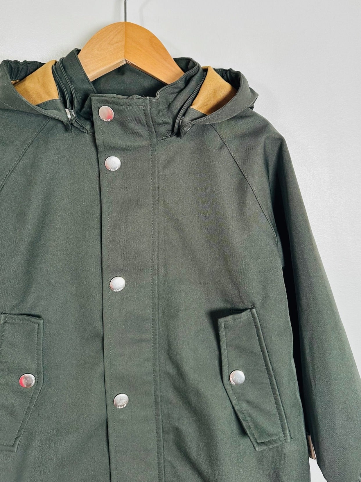 An olive green hooded raincoat with silver snap buttons, hanging on a wooden hanger against a plain light gray wall.