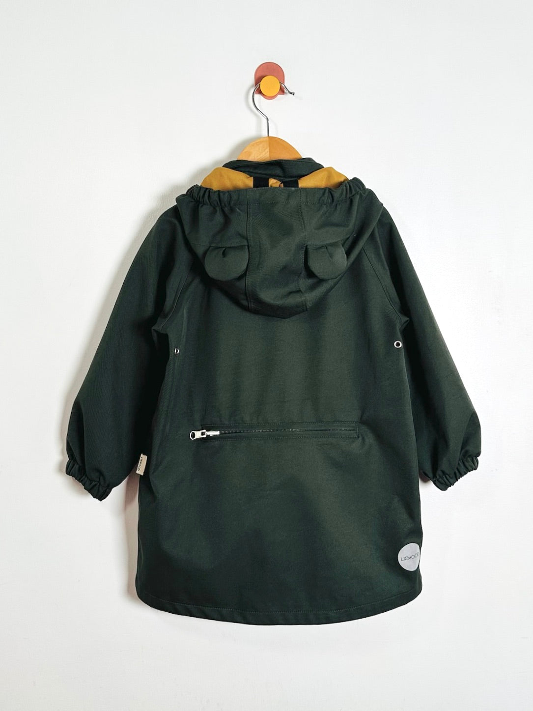 A dark green hooded raincoat hanging on a wooden hanger against a plain white wall. The coat features a hood, elasticized cuffs, a front zippered pocket, and a small circular white label near the lower right hem.