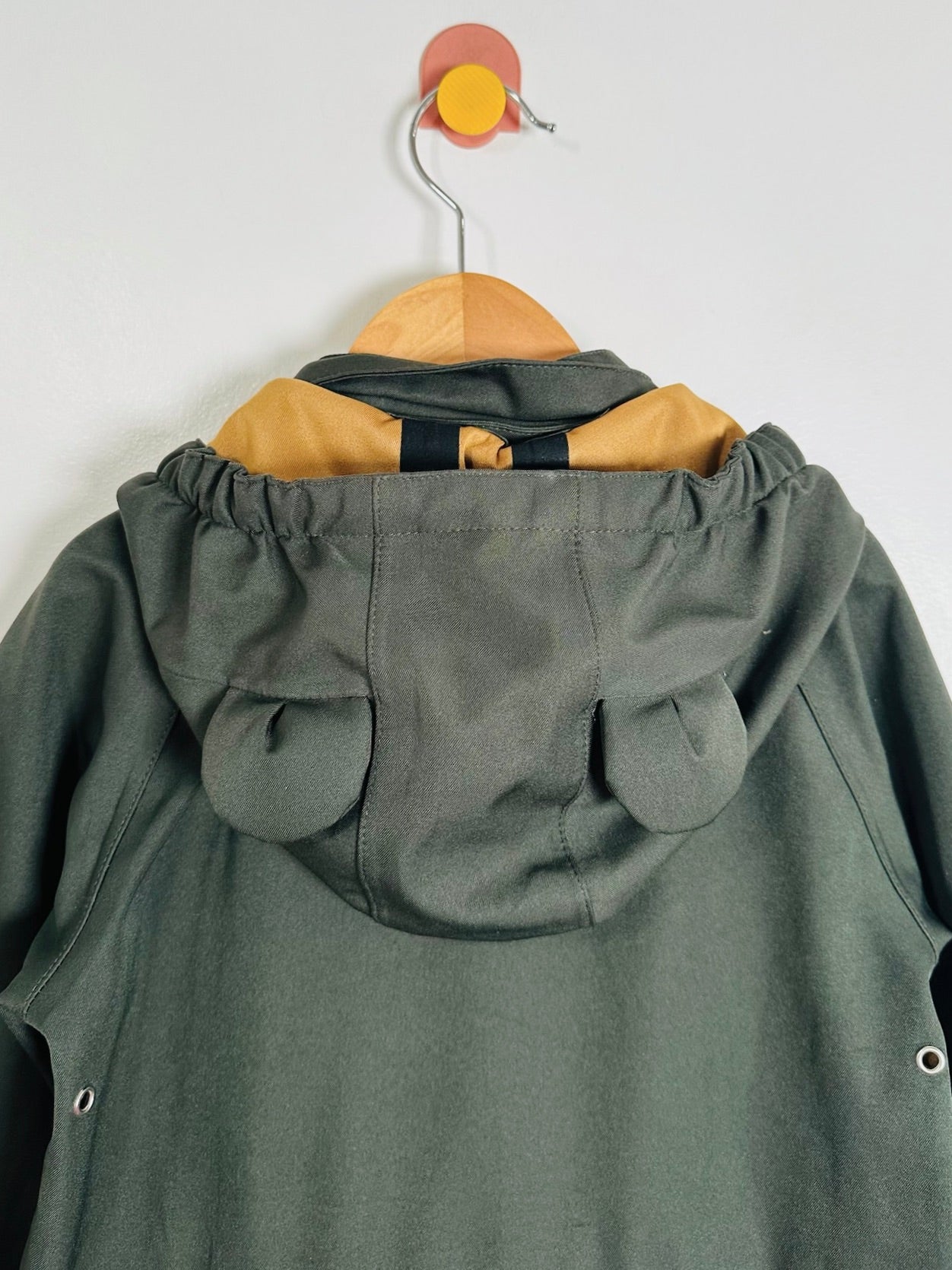 An olive green hooded jacket with a tan inner hood lining and black drawstrings, hanging on a wooden hanger with a wall hook.