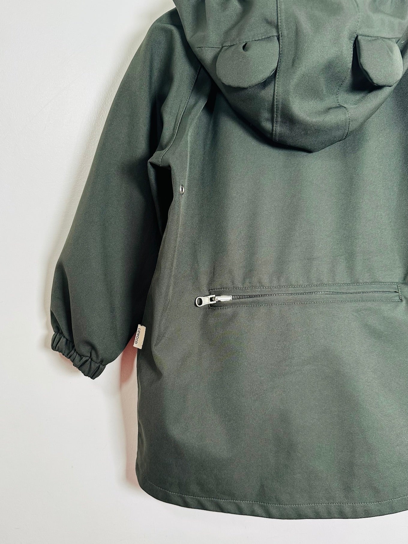 An olive green hooded jacket shown from the back, featuring a zippered pocket and elasticized cuffs, photographed against a plain white background.