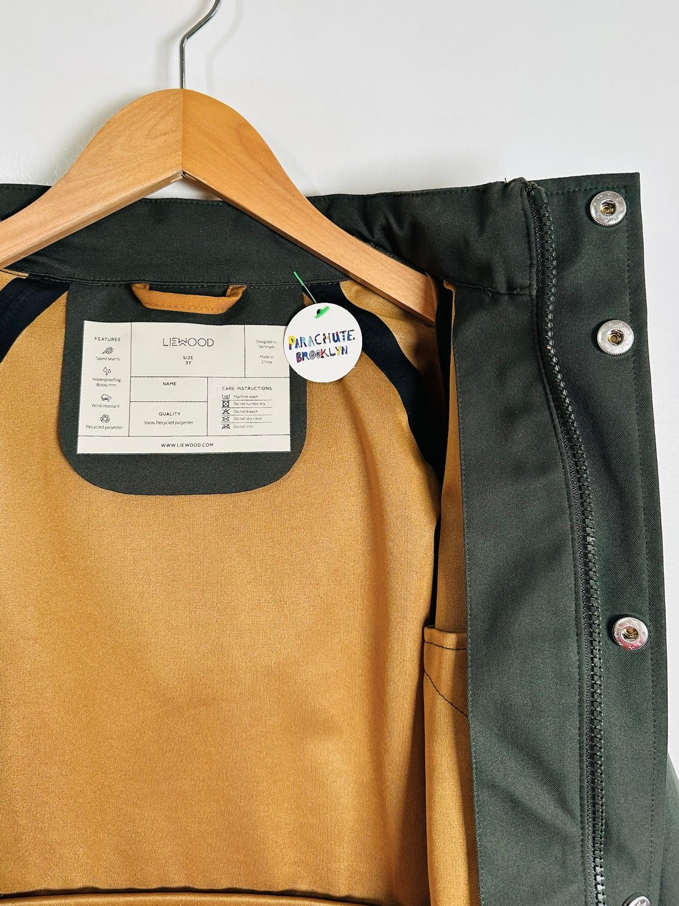 A dark green and tan jacket from the brand Lethood, shown hanging on a wooden hanger against a plain white wall. The jacket is open to display its interior lining and a side pocket with metal snap buttons.