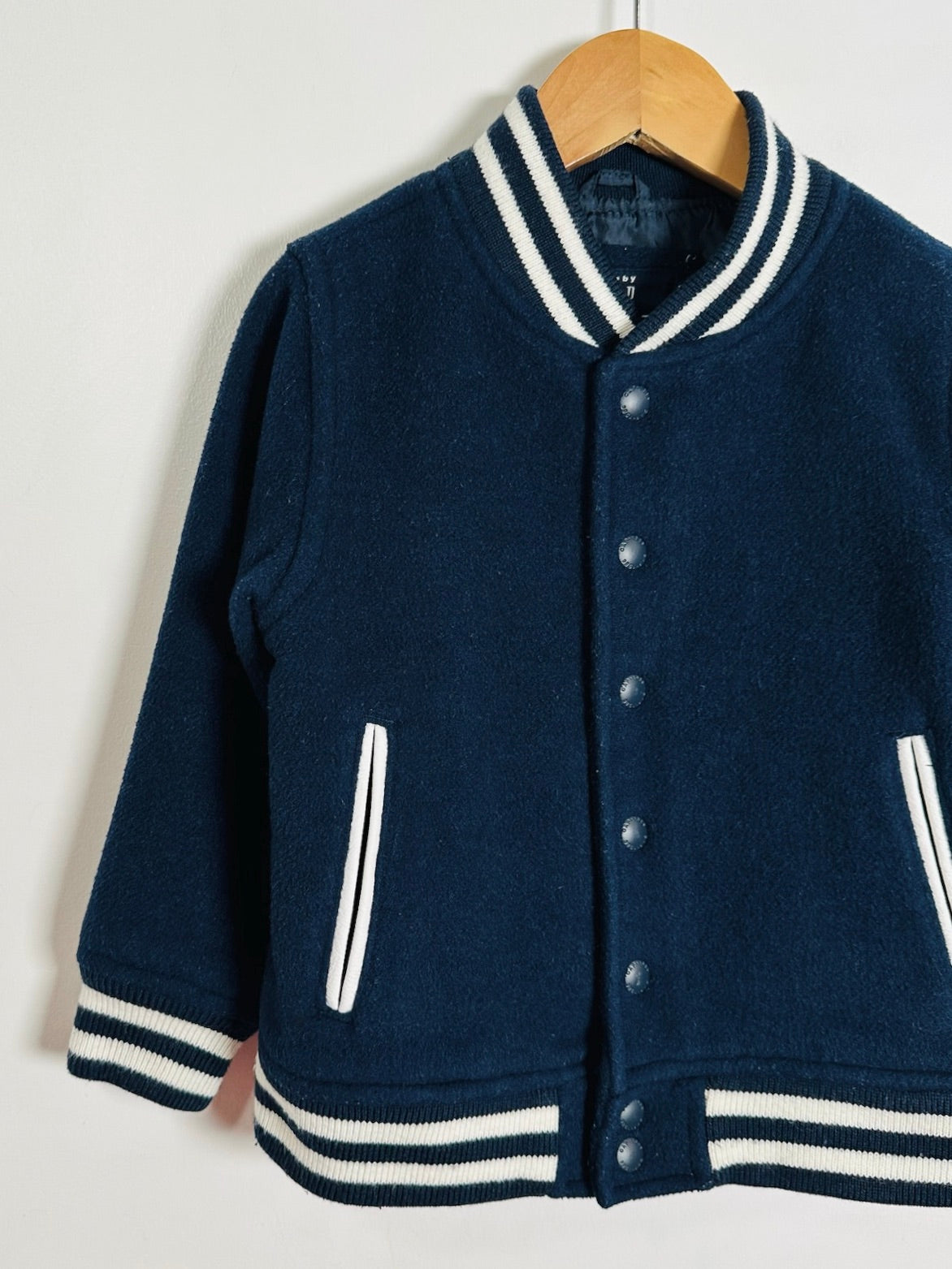 A navy blue wool-blend varsity jacket with white striped ribbed trim, hanging on a wooden hanger against a plain off-white wall.