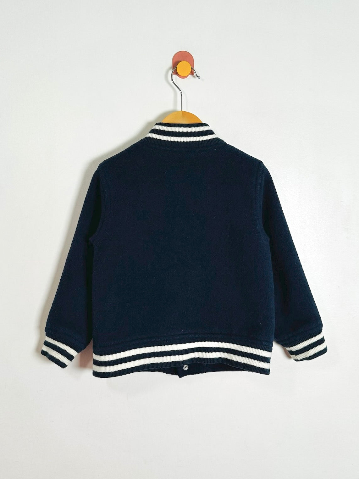 A navy varsity jacket with white striped ribbed trim on the collar, cuffs, and hem, hanging on a wooden hanger against a plain white wall.