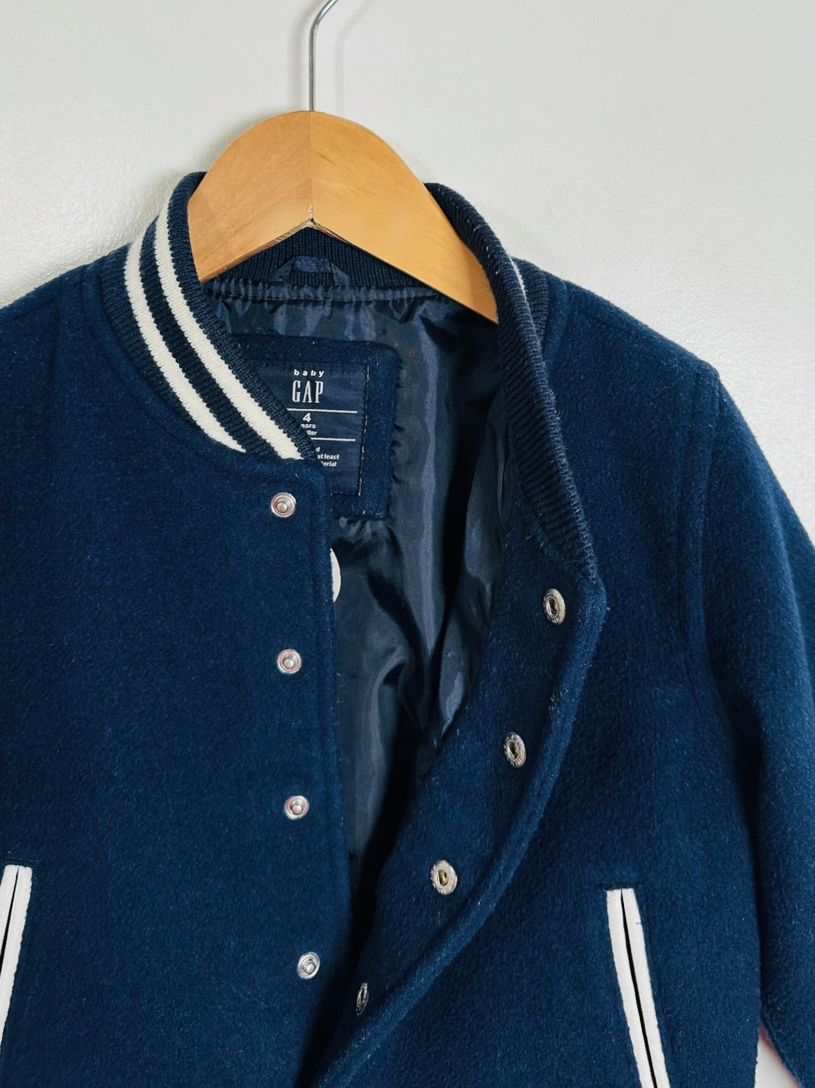 A navy blue knit varsity-style jacket hanging on a wooden hanger against a plain light wall. The jacket has white striped ribbed trim on the collar and sleeve cuffs, silver snap buttons, and a visible GAP neck label.