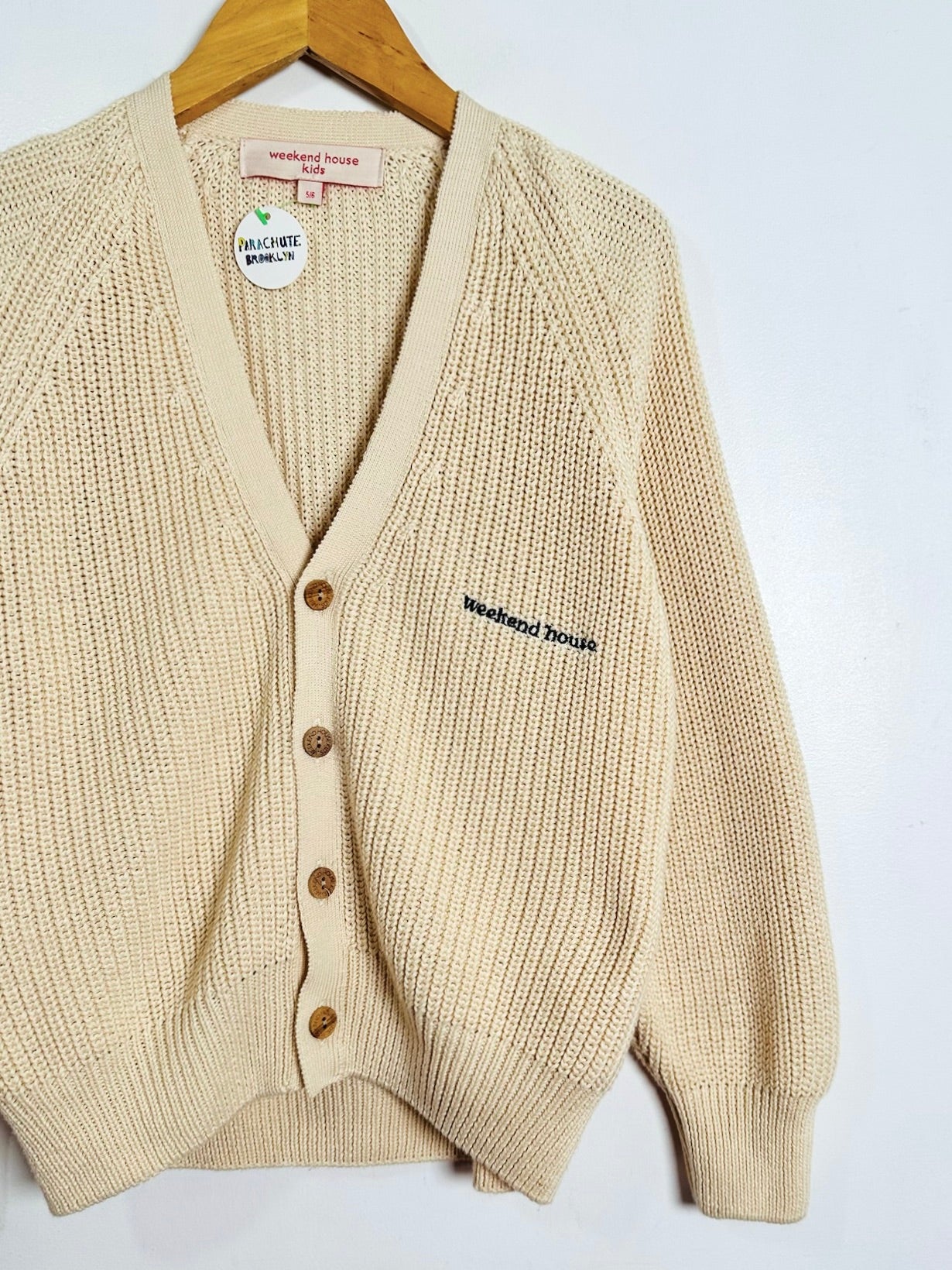 A cream-colored ribbed knit cardigan sweater with brown buttons, hanging on a wooden hanger against a plain white background. The cardigan features 'Weekend Beach' embroidered on the left chest.