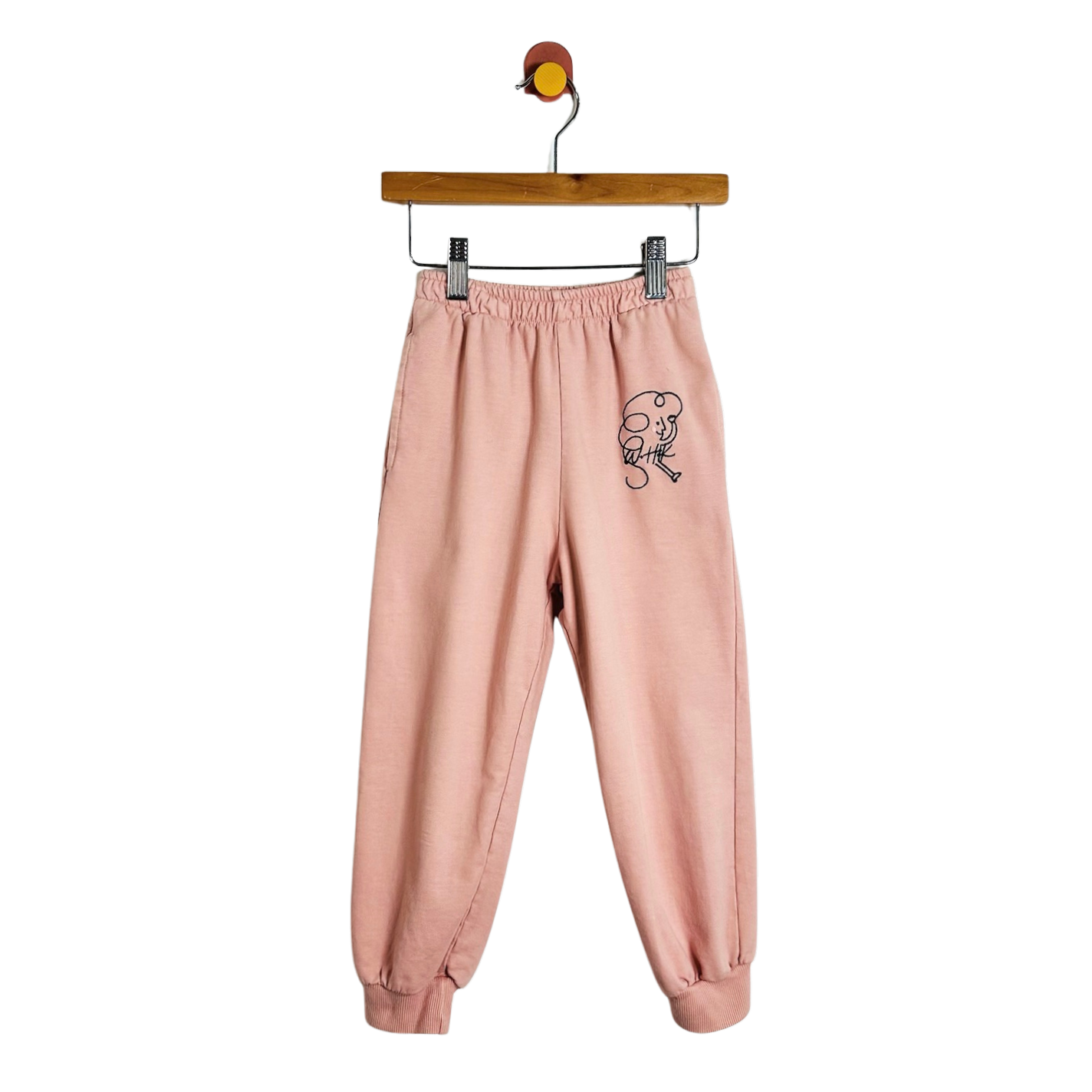 A pair of light pink sweatpants with a small black cartoon graphic on the left thigh, hanging from a wooden hanger against a plain white background.