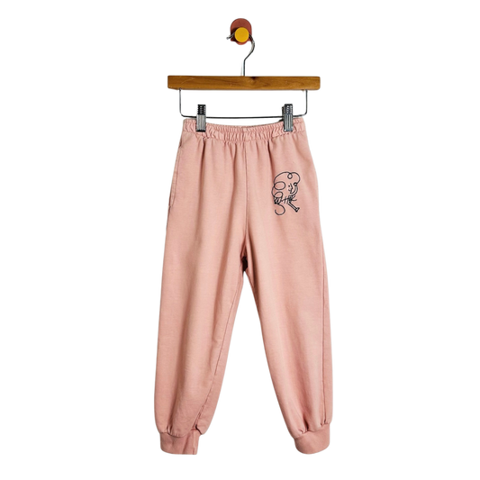A pair of light pink sweatpants with a small black cartoon graphic on the left thigh, hanging from a wooden hanger against a plain white background.