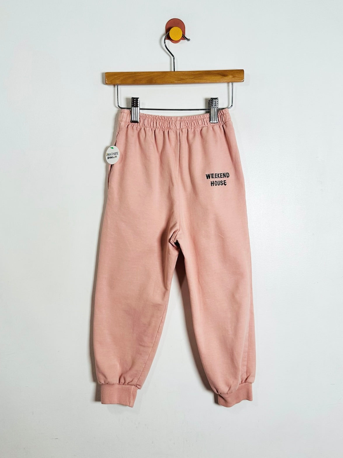 A pair of dusty pink sweatpants with 'WEEKEND HOUSE' printed in black on the left thigh, hanging from a wooden hanger against a plain white wall.
