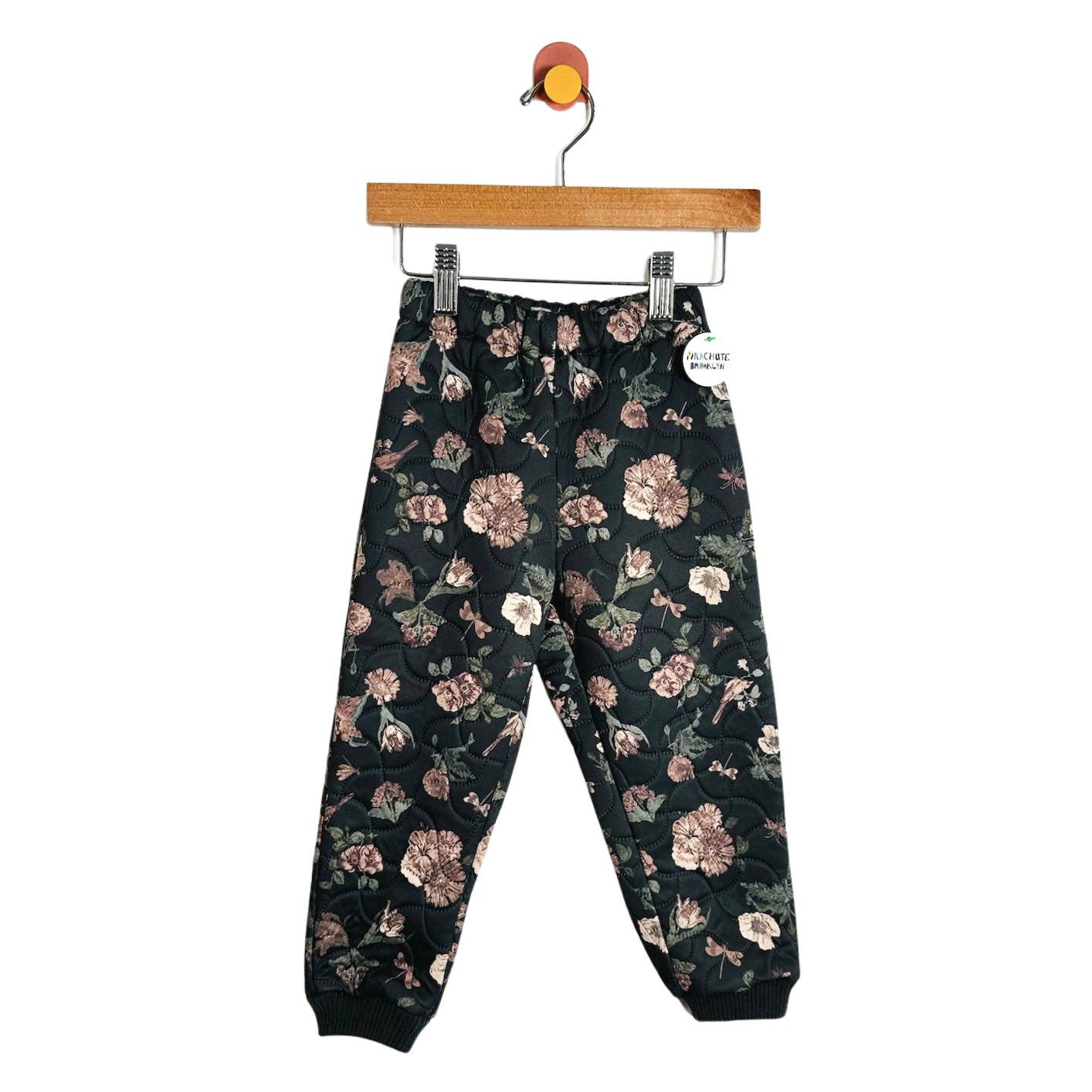 A pair of children's jogger pants with a dark green floral pattern, hanging from a wooden hanger against a plain white background.