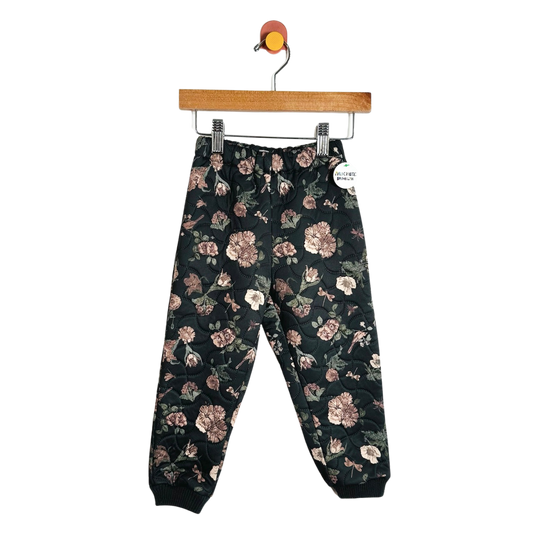 A pair of children's jogger pants with a dark green floral pattern, hanging from a wooden hanger against a plain white background.
