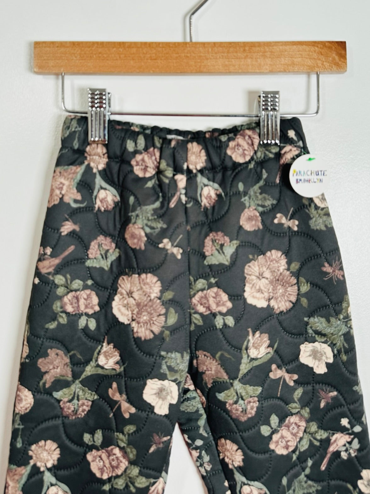 A pair of quilted pants with a floral print hanging from a wooden hanger. The pants feature a dark charcoal background with pink, beige, and green floral motifs and are shown against a plain white wall.