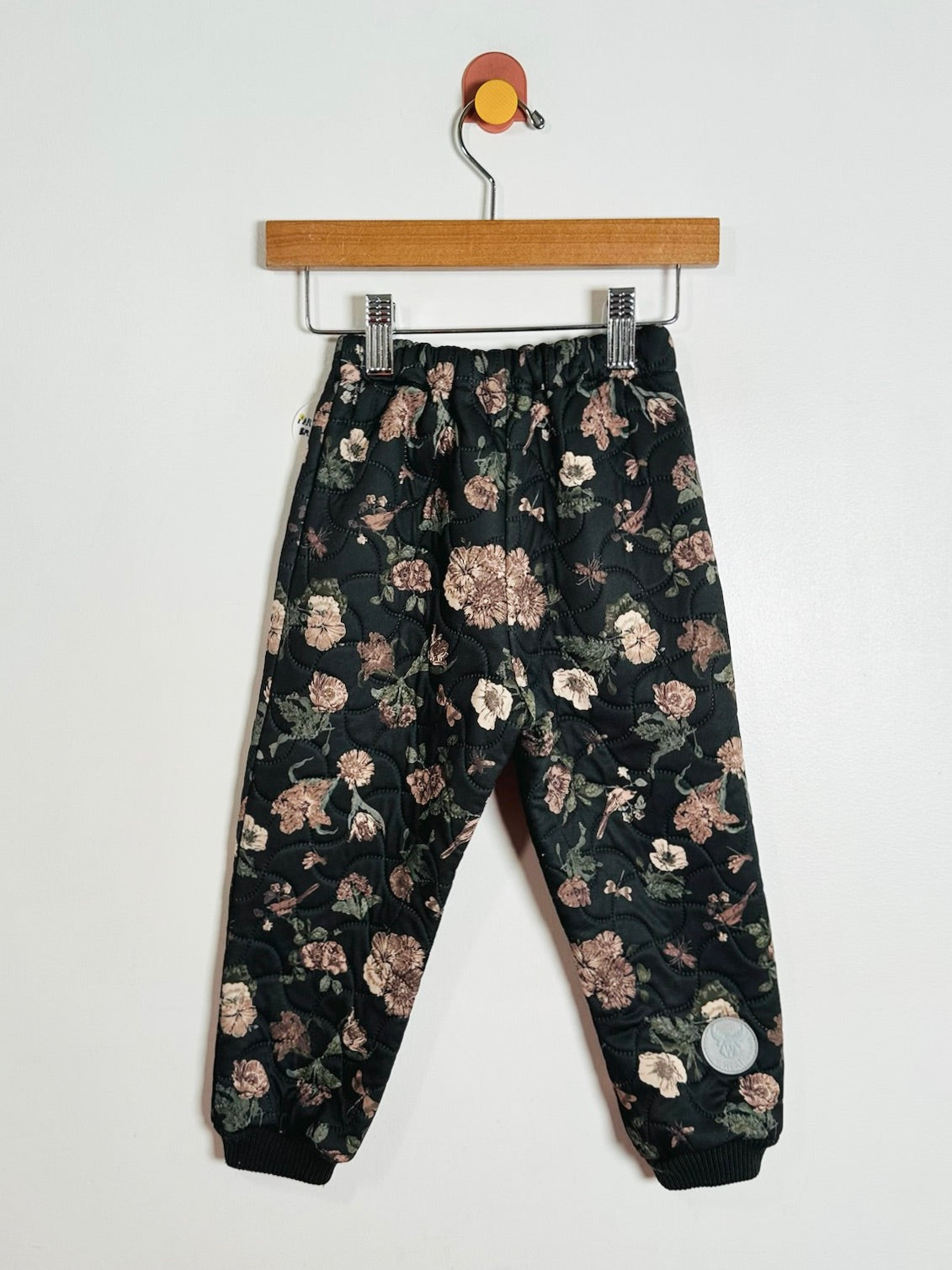A pair of black children's pants with an all-over floral print in muted pink, beige, and green, hanging from a wooden hanger against a plain white wall.