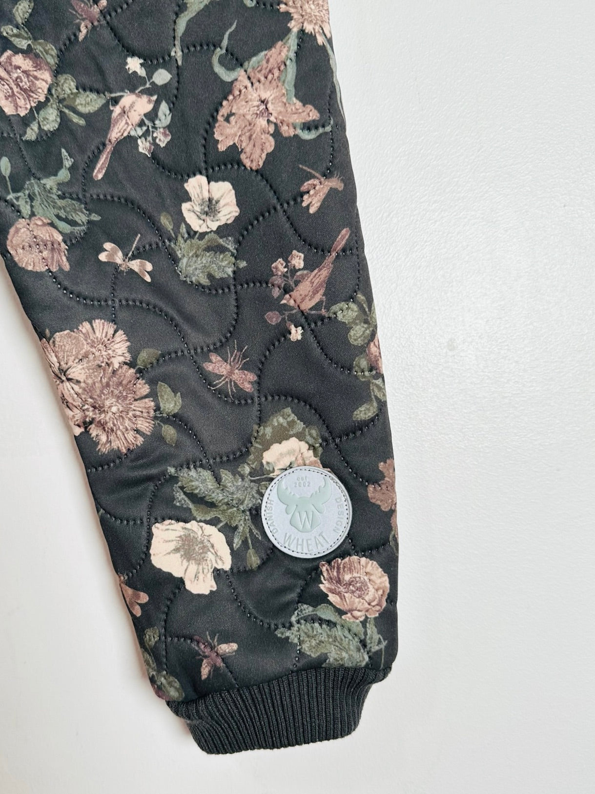 A close-up of quilted pants featuring a dark charcoal background with a floral and bird pattern in muted pink, beige, and sage green. A circular light blue-gray brand patch is sewn onto the leg near the cuff.