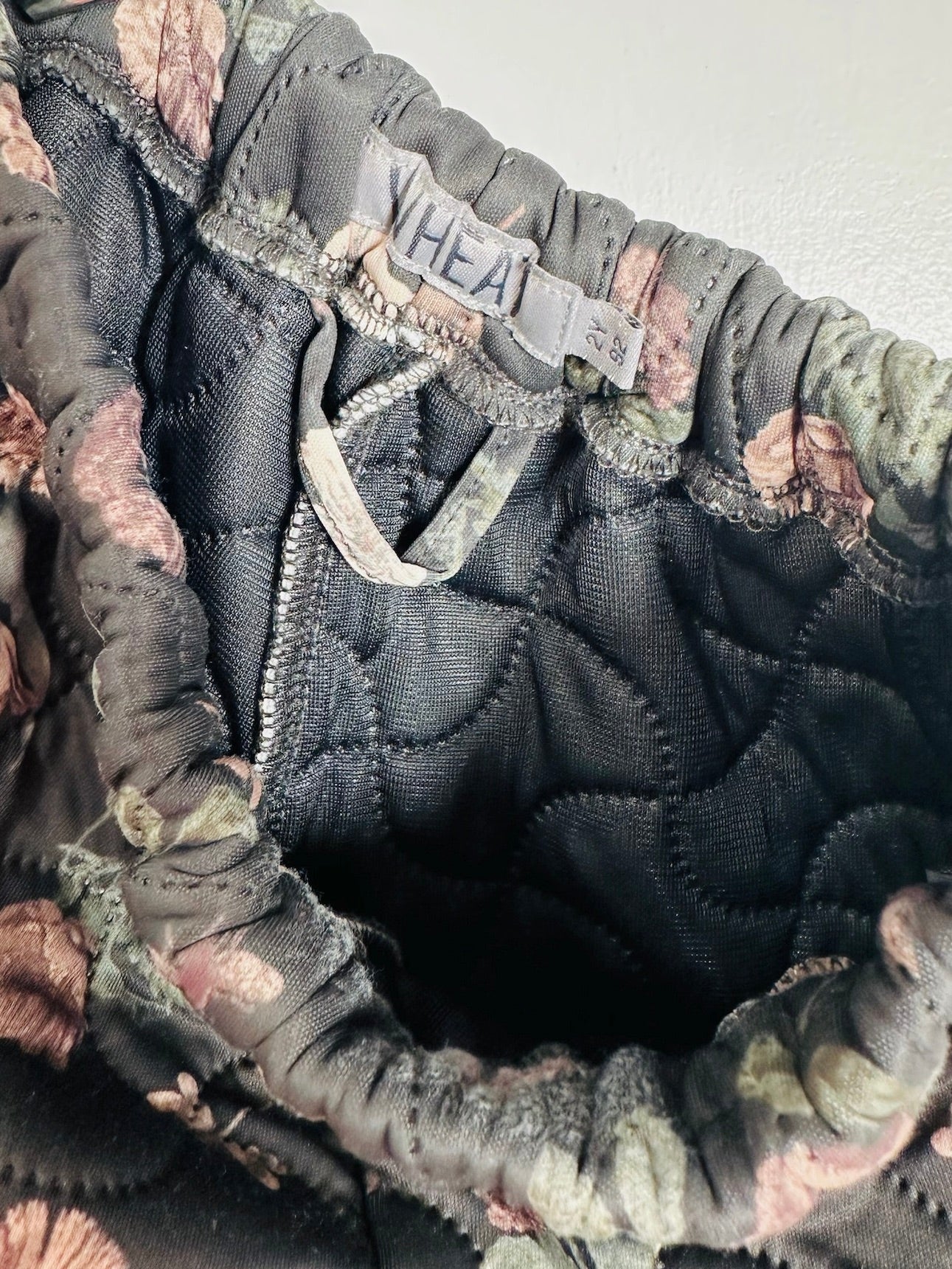 Close-up of a quilted garment opening showing a dark charcoal gray interior with floral trim in muted pink, green, and brown tones. A sewn-in label reads 'WHEAT'.