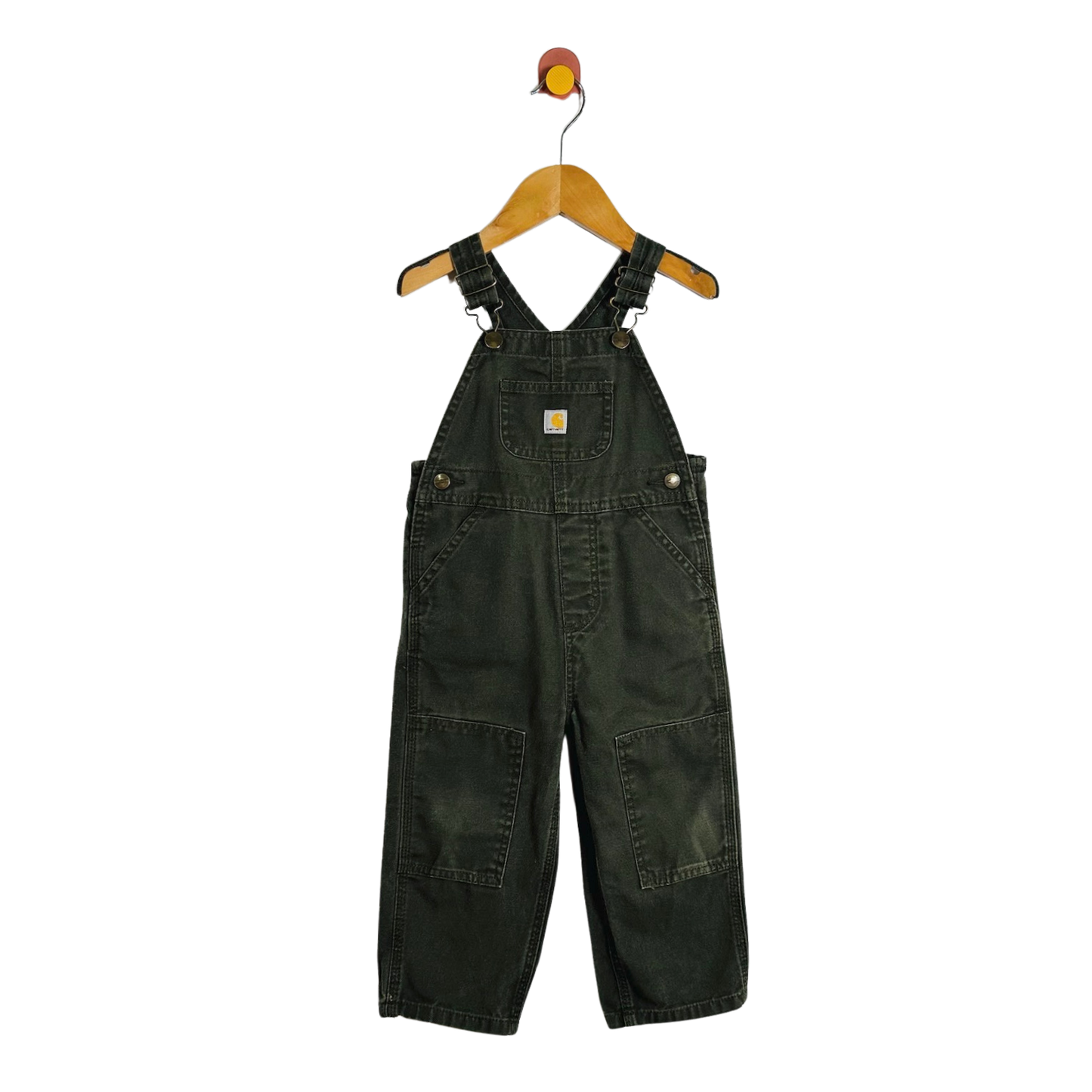 A pair of dark green denim baby overalls hanging on a wooden hanger with a red and yellow hook against a plain white background.
