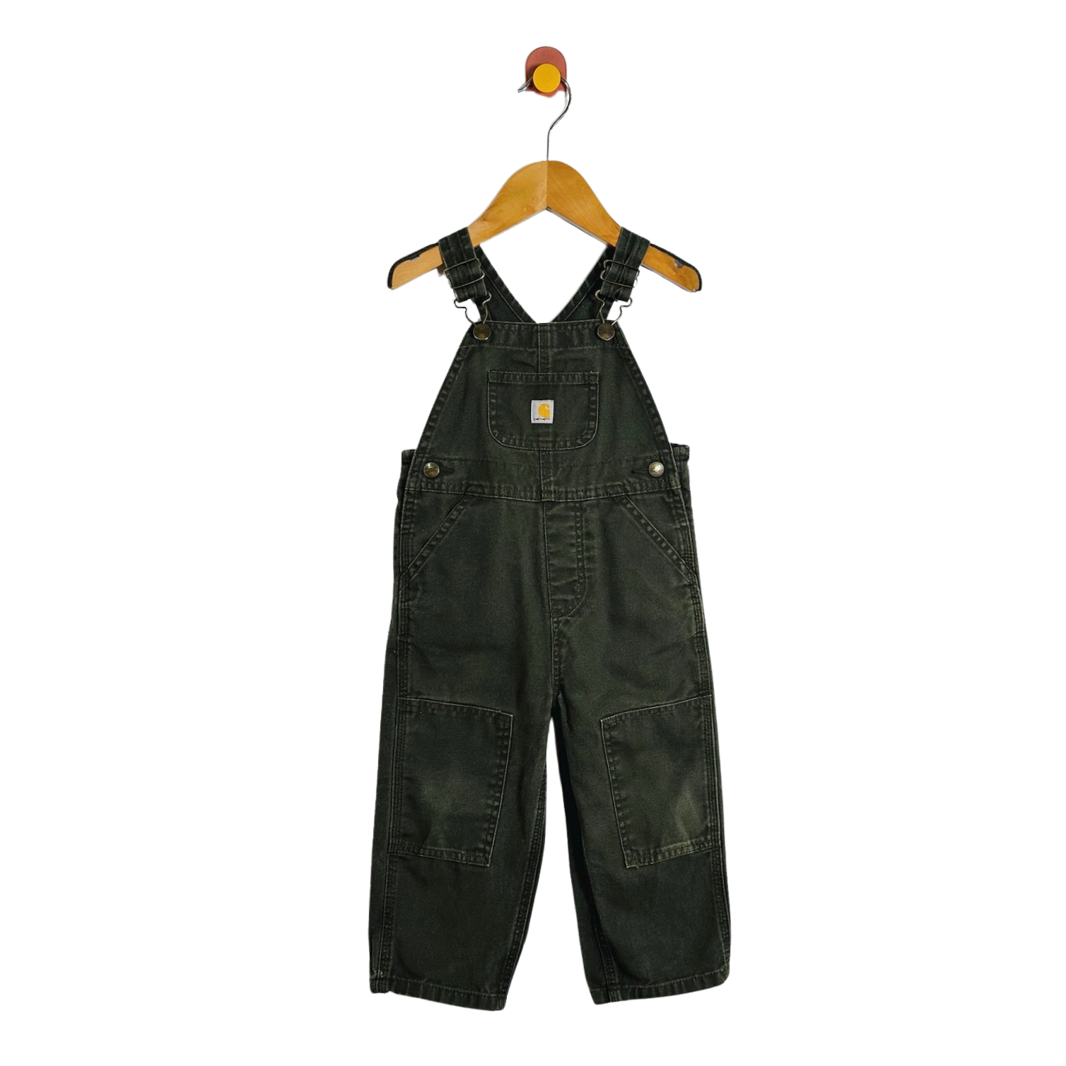 A pair of dark green denim baby overalls hanging on a wooden hanger with a red and yellow hook against a plain white background.