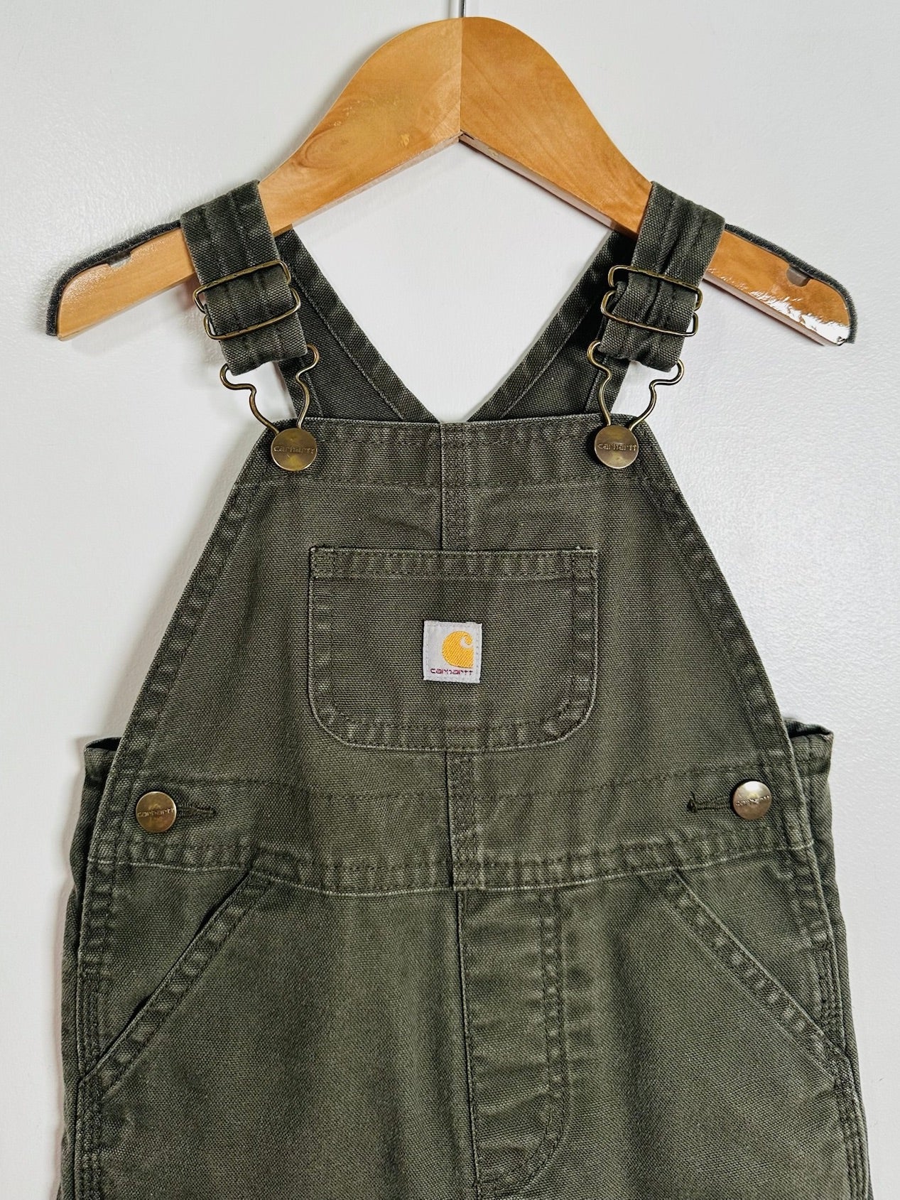 An olive green Carhartt denim bib overalls hanging on a wooden hanger against a white wall. The overalls feature adjustable shoulder straps with metal snap buttons, a front patch pocket with a visible Carhartt logo, and side pockets.