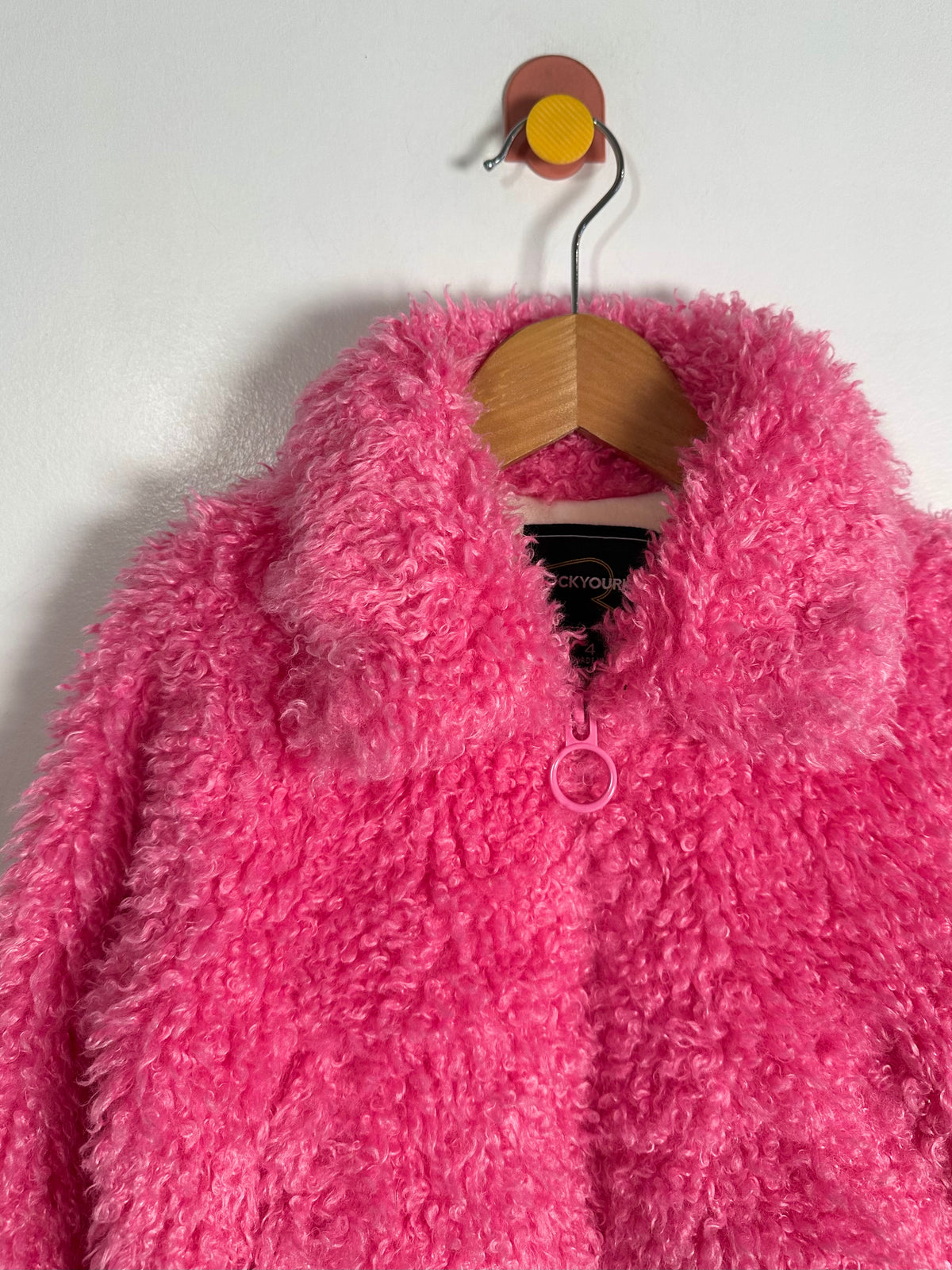 A bright pink fuzzy sherpa fleece jacket hanging on a wooden hanger against a plain white wall.