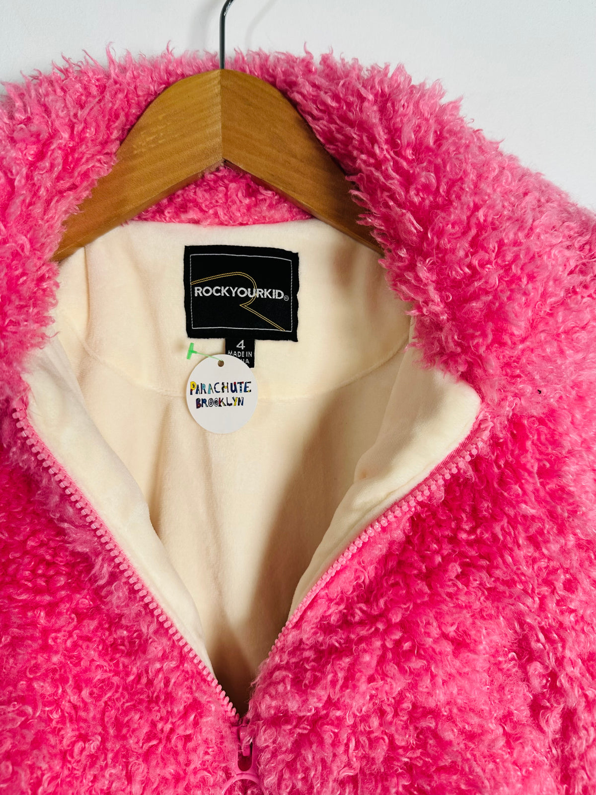 A close-up of a bright pink fuzzy sherpa zip jacket hanging on a wooden hanger against a white background. The inner label reads 'ROCKYOURKID' and a hangtag 