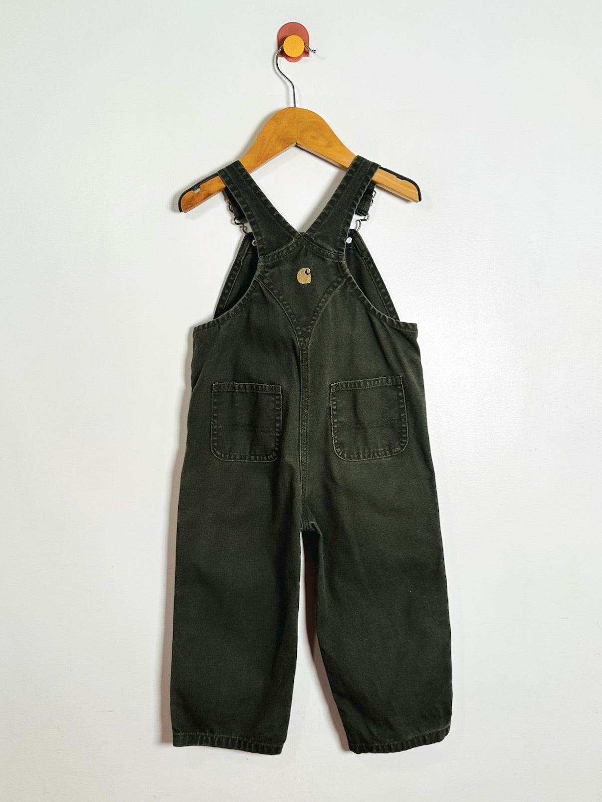 A pair of dark olive green bib overalls hanging from a wooden hanger against a plain white wall. The overalls feature adjustable shoulder straps, a small gold-tone metal logo button on the chest, and two front patch pockets.