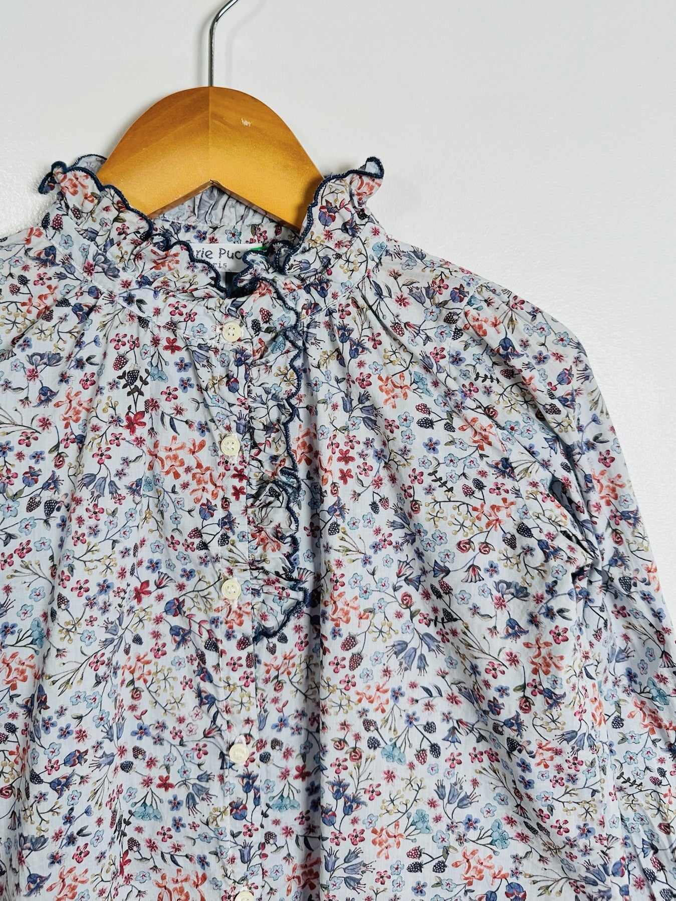 A close-up of a long-sleeve blouse with a ruffled high neck and an all-over small floral print, hanging on a wooden hanger against a plain light background.