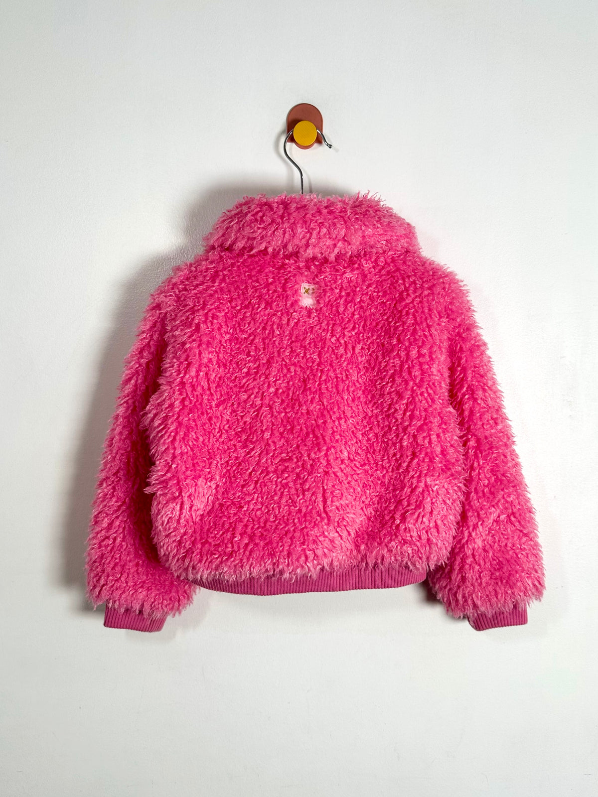 A bright pink fuzzy teddy fleece jacket hanging on a metal hanger against a plain white wall.