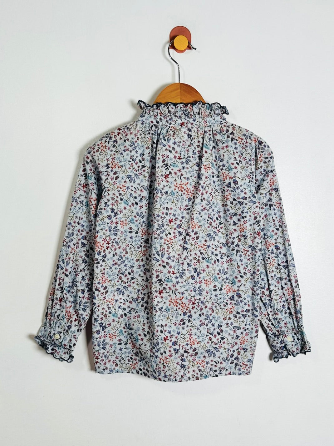 A long-sleeve blouse with a dense small floral print in muted blue, red, and pink tones on a light gray base, hanging on a wooden hanger against a plain off-white wall.
