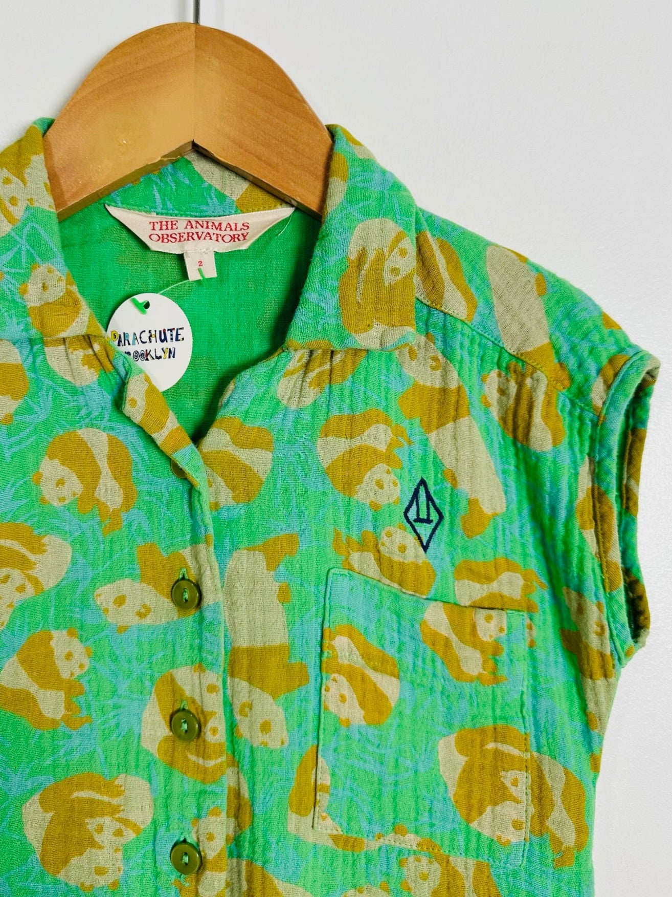 A green short-sleeve button-up shirt with a yellow and beige animal print, hanging on a wooden hanger against a plain white wall. The shirt features a chest pocket with a small embroidered logo and a visible 'The Animal & Movements' brand label.
