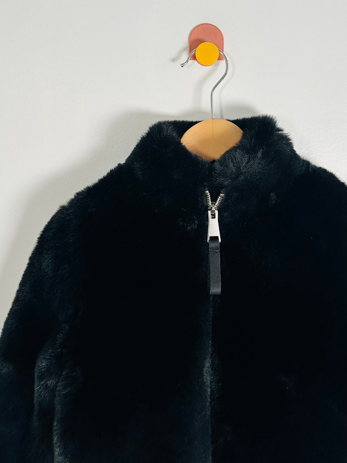 A black faux fur jacket with a high collar and a silver zipper, hanging on a wooden hanger against a plain white wall.