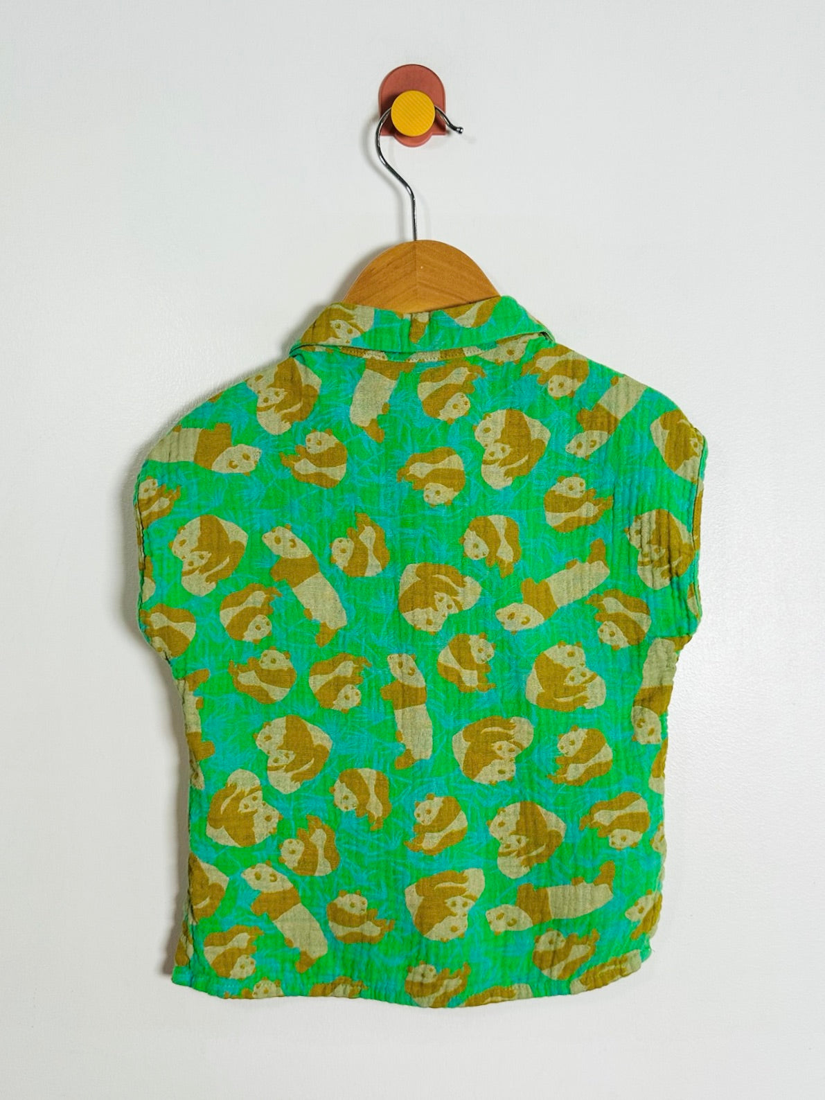 A green sleeveless collared top with an all-over tan monkey print, hanging on a wooden hanger from a wall hook against a plain white wall.